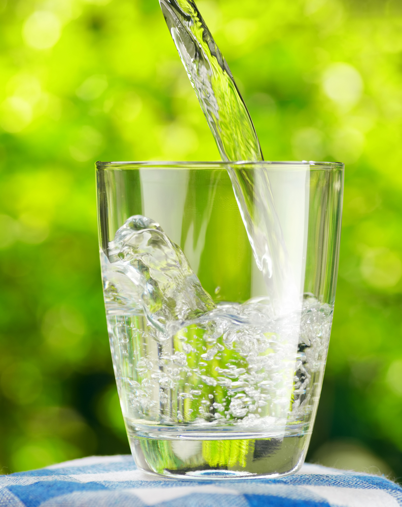 Clear glass being filled with water on blue checkered blanket with bright, sunny background of green trees