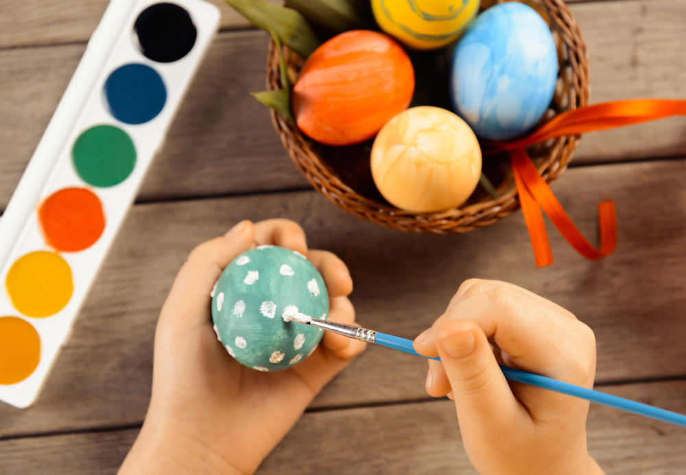 Child painting Easter eggs with brush, paint and Easter eggs in basket in background on wooden table