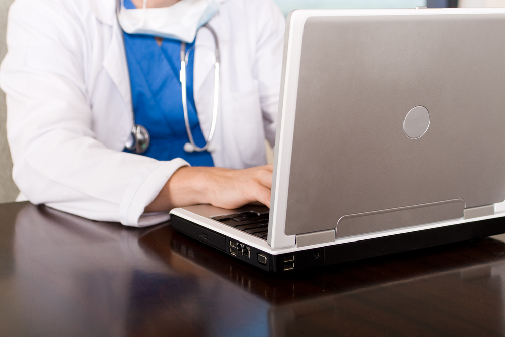 Doctor in scrubs and white coat with stethoscope and face mask working on laptop sitting on top of wooden table