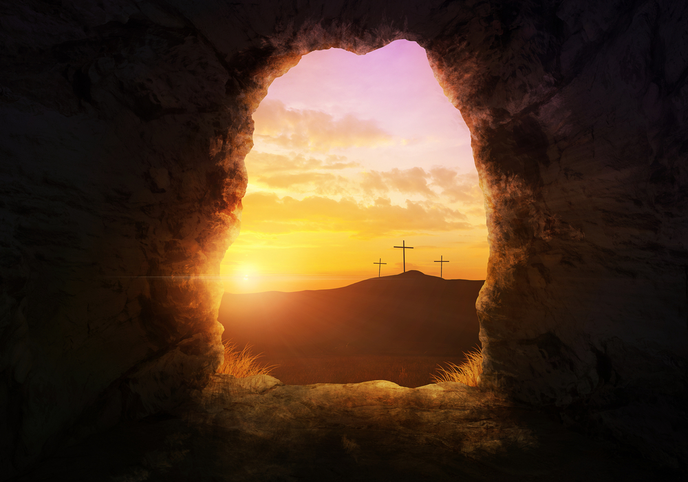 Empty tomb of resurrected Jesus open to a view of a sunrise and three crosses on a hill