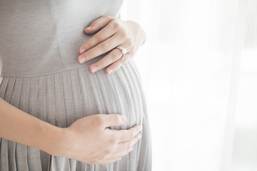 Pregnant Woman in Gray Dress Holding Baby Bump with White Curtains in Background