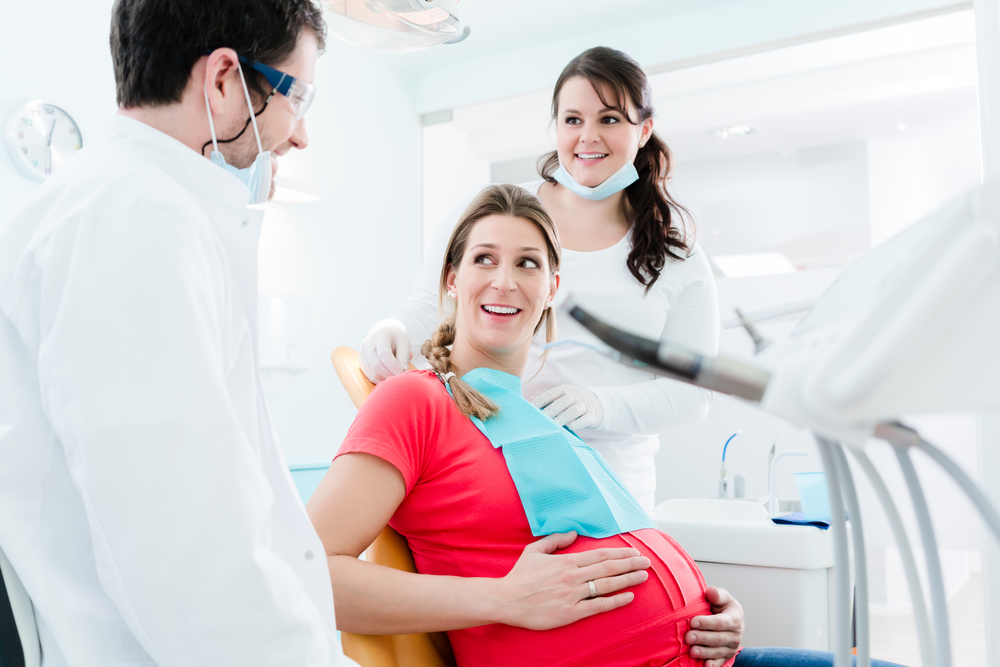 Pregnant woman in pink shirt holding baby bump in dental examination chair with dental hygienist in white putting on protective apron on patient and dentist in white coat talking to patient with white walls and equipment