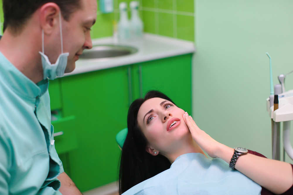 Woman with toothache in dental chair next to dental equipment holding side of face explaining dental problem to dentist with mask, green sink and backsplash in background