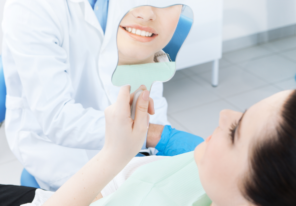 Woman patient looking at healthy teeth in mirror with dentist in background