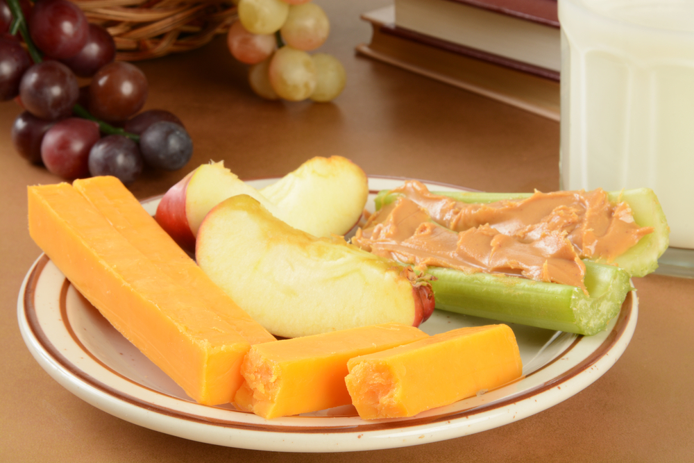 Plate of cheddar cheese sticks apple slices and celery with peanut butter, with grapes and a glass of milk in the background