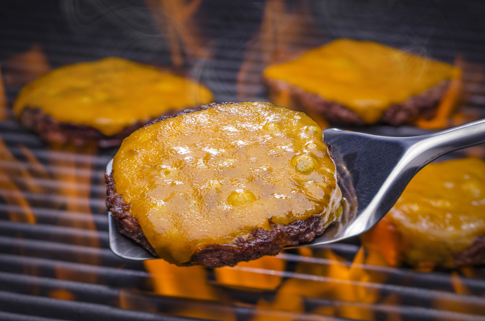 Spatula holding cheeseburger of grill with flames cooking 3 other patties with cheese