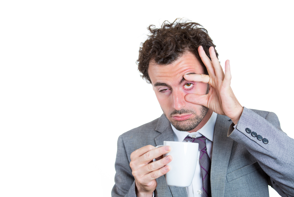 Tired man holding a cup of coffee with one hand and trying to hold his eye open with the other hand