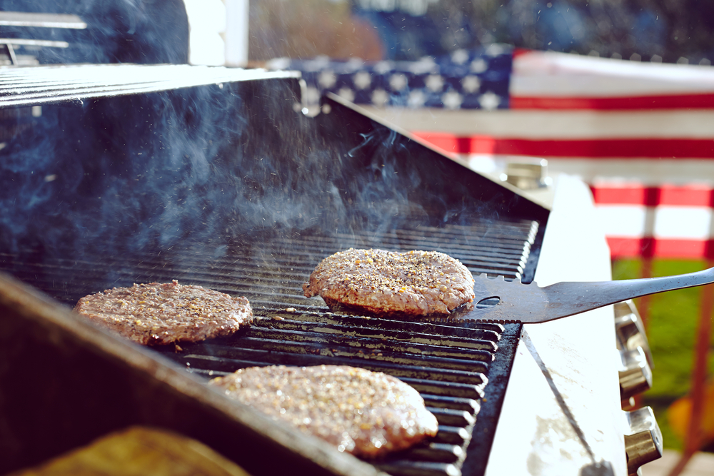 Spatula holding burger patty above grill with other paties with American flag in background