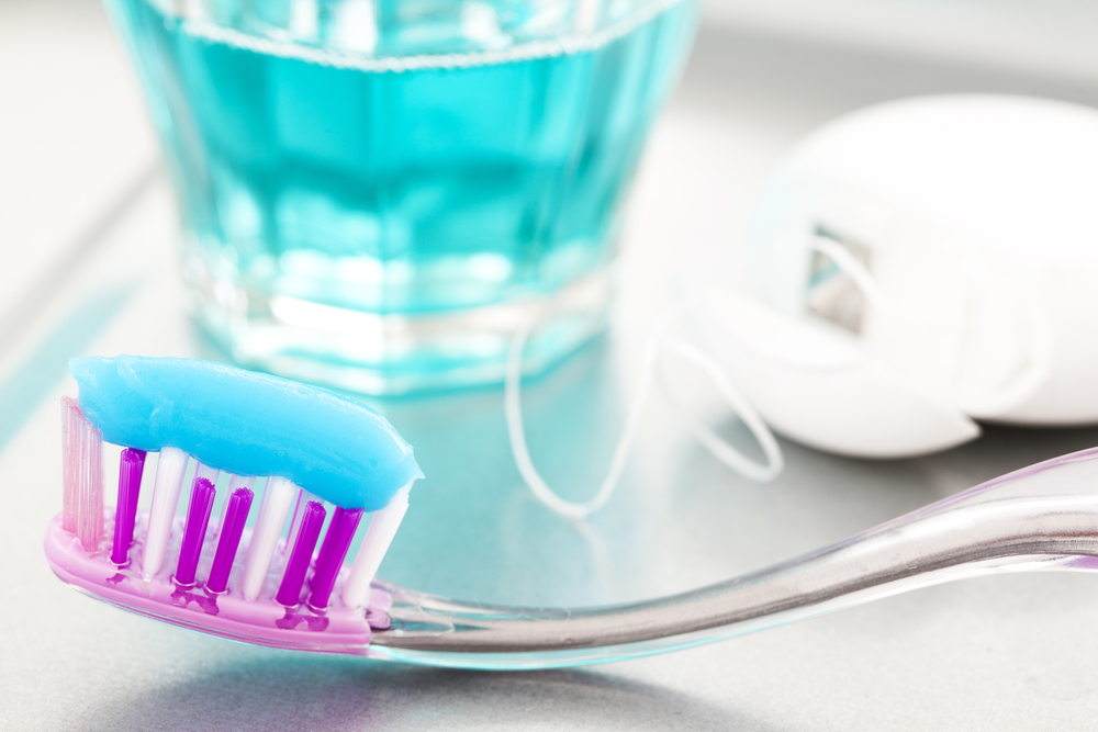 Toothpaste on purple and white bristes toothbrush with glass of mouthwash and container of floss in background