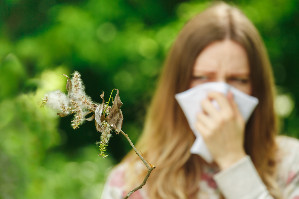 girl suffering from summertime sinuses is holding a tissue over nose staring at a pollen spreading plant 