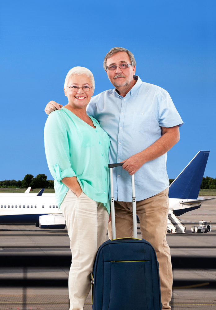 Husband and wife with carry-on suitcase standing in airport with airplane in background