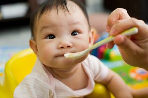 Six month old South East Asian Chinese baby girl sitting in a yellow seat, being spoon fed